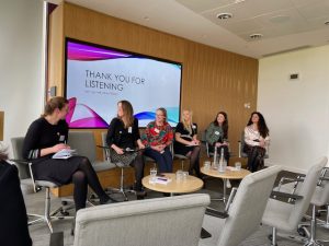 Photo taken at the event showing the five panellists and keynote speaker Holly Redman at the front of the room, listening to a question from the chair Parminder Lally (who is to one side, not visible)
