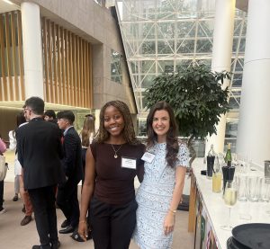 A Black woman and a white woman posing together and smiling for the camera at a networking event in an office reception room, with other attendees behind them and a drinks table to one side