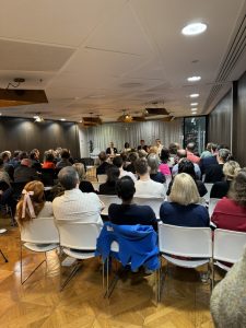 photo of the event taken from the back of the audience: rows of people in chairs with their backs to the camera and facing the panel of 4 speakers who are sat at the front of the room