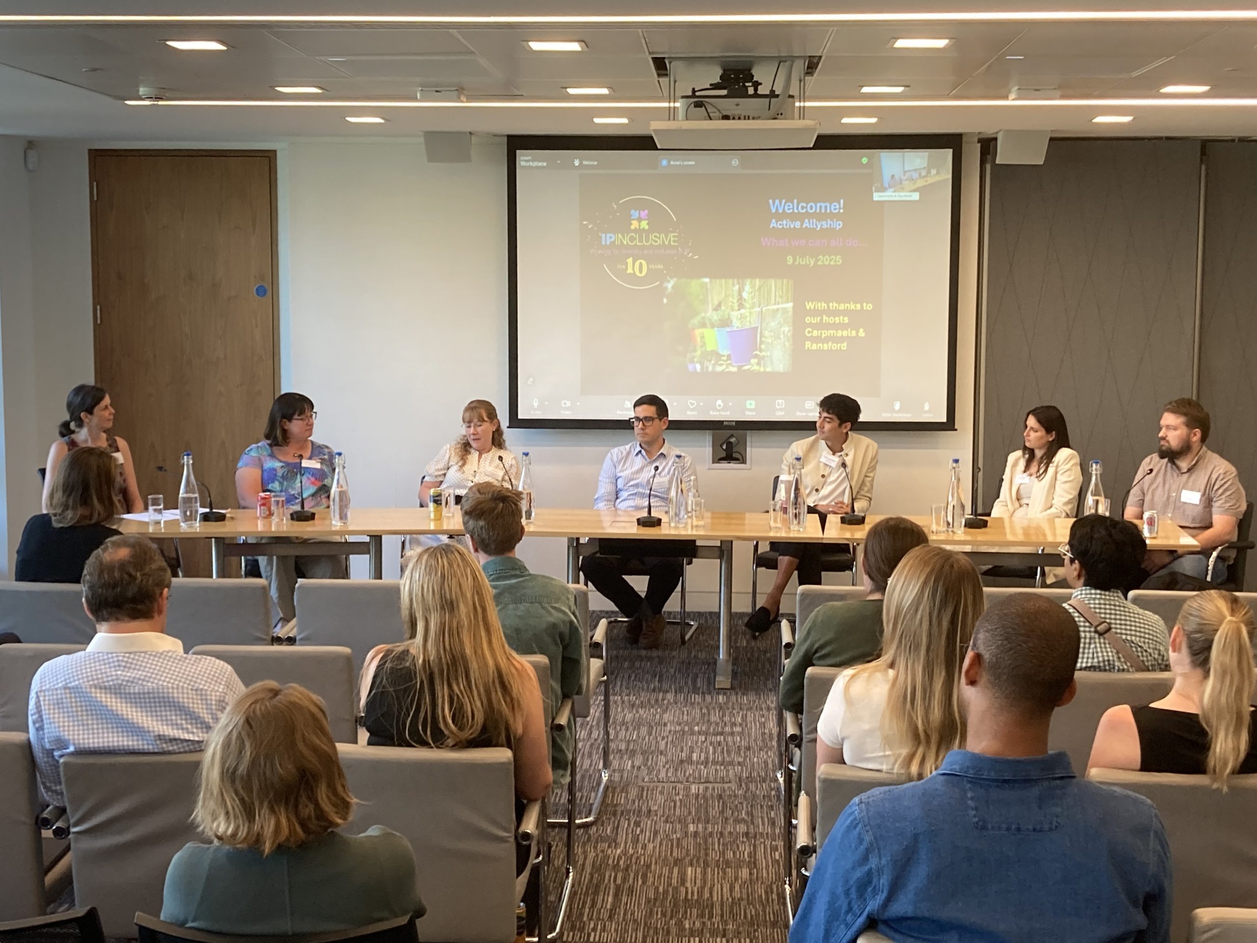 Photo taken at the event showing the chair person and six panellists sitting at a long table, with some audience members observing the discussion. 
