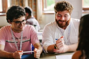 A photo of two students working on a problem. both are smiling, one has a tape measure around his neck and is holding a ruler, the other is holding a pen.